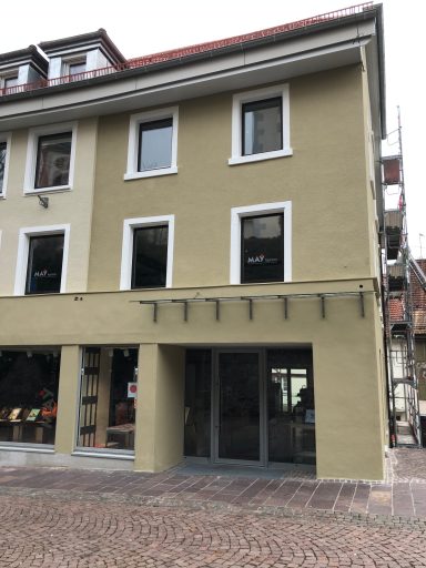 Helles Gebäude mit mehreren Fenstern und einem Eingangsbereich, im Stadtbild eingebettet.