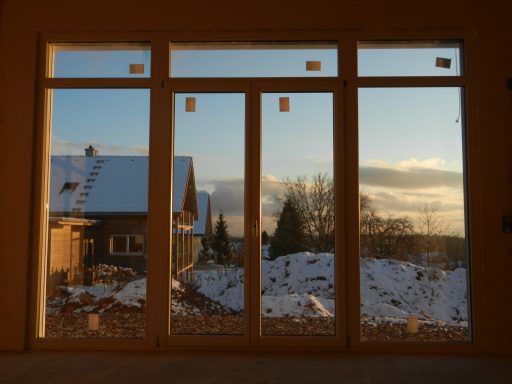 Blick durch große Fenster auf verschneite Landschaft und ein Haus im Hintergrund bei Sonnenuntergang.